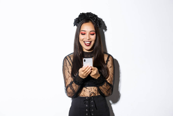 Nadšený usmívající se žena při pohledu na mobilní telefon potěšen, na sobě gotické krajkové šaty pro halloween party, stojí nad bílým pozadím - Fotografie, Obrázek