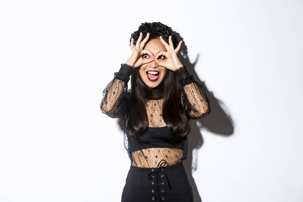 Amazed beautiful asian woman in halloween costume looking through finger glasses with excited happy face, standing over white background wearing gothic witch dress - Photo, Image