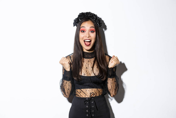 Изображение счастливой азиатской готической девушки в праздничном платье на Хэллоуин, радующейся победе, торжествующей и говорящей "да", стоящей на белом фоне и празднующей победу - Фото, изображение