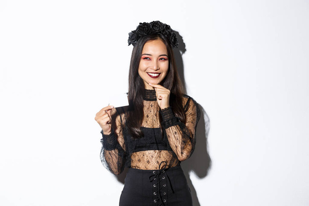 Imagen de mujer asiática sonriente reflexiva en traje de Halloween tienen una idea, la celebración de la tarjeta de crédito y buscando astucia en la esquina superior izquierda - Foto, imagen