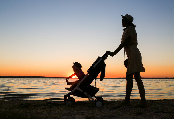 Gün batımında nehrin kenarında bebek arabasında bir kadın silueti. - Fotoğraf, Görsel
