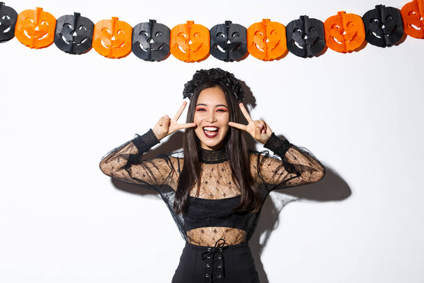 Cute happy asian woman enjoying halloween party, showing peace gesture and smiling, wearing witch costume, standing against pumpkin banners decoration - Фото, изображение