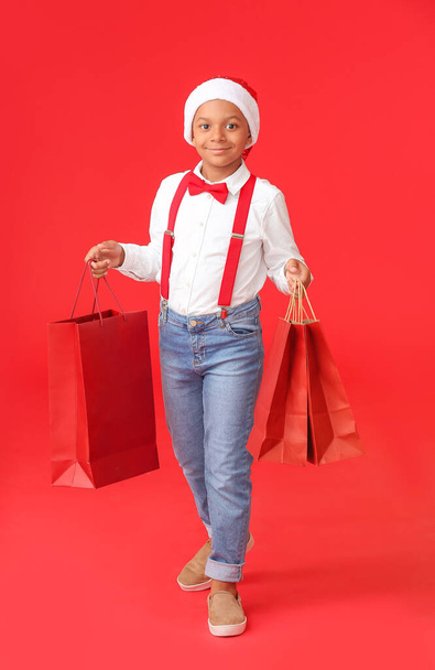 Cute African-American boy in Santa hat and with shopping bags on color background - Zdjęcie, obraz