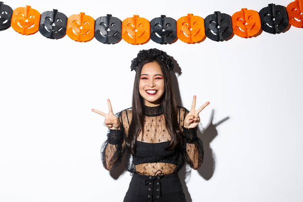Cute happy asian woman enjoying halloween party, showing peace gesture and smiling, wearing witch costume, standing against pumpkin banners decoration - Фото, изображение
