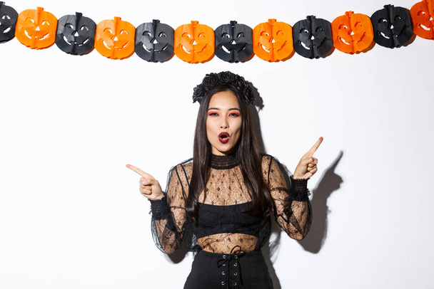 Joyeuse femme asiatique en robe de dentelle gothique célébrant Halloween, pointant du doigt sur le côté, debout sur fond blanc avec des banderoles de fête citrouille - Photo, image