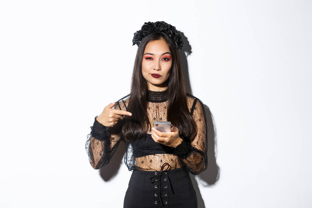 Sassy bella donna asiatica in abito gotico e ghirlanda nera che punta il dito al telefono cellulare, mostrando qualcosa su Halloween - Foto, immagini
