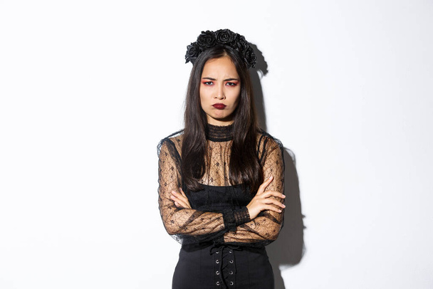 Bild von wütenden und beleidigten asiatischen Mädchen, die sich über irgendetwas beschweren, die Arme verschränken und schmollen, beleidigt oder eifersüchtig gucken, vor weißem Hintergrund im Halloween-Party-Kleid stehen - Foto, Bild