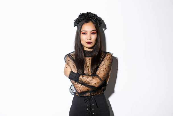 Young elegant woman in stylish gothic dress and wreath, embracing her body and looking sassy at camera, wearing halloween costume of undead widow, standing over white background - Photo, Image