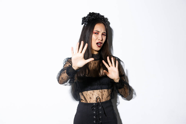 Image of bothered and annoyed asian woman in elegant gothic dress raising hands defensive, grimacing from camera flesh, asking to stop taking pictures of her, standing white background - Photo, Image
