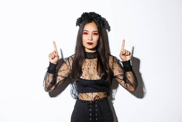 Sassy piękna azjatycka kobieta w czarnym gotyckim stroju, nosząc strój czarownicy na halloween i wskazując palce w górę, pokazując swoje logo lub baner na pustym białym tle, białe tło - Zdjęcie, obraz