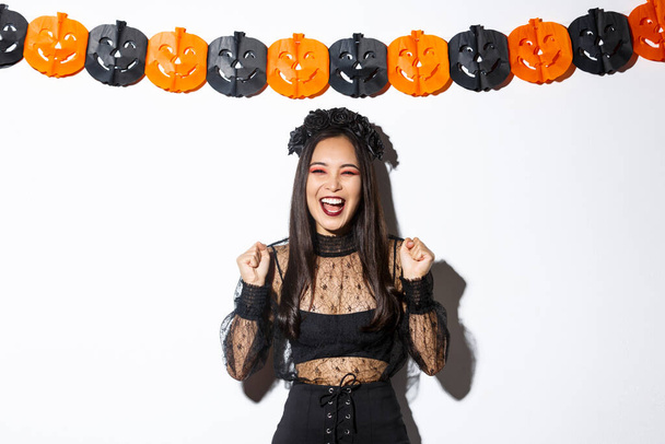 Femme asiatique souriante insouciante en costume de sorcière profitant de fête d'Halloween, dansant et se réjouissant, debout sur fond blanc avec décoration de banderoles de citrouille - Photo, image