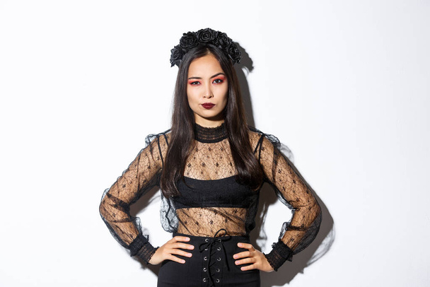 Atractiva mujer asiática en traje de halloween buscando decepcionado y escéptico. Mujer en vestido de encaje negro y corona con aspecto arrogante, truco o trato en traje de bruja, de pie fondo blanco - Foto, Imagen