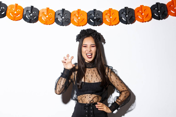 Image of girl in wicked witch costume acting evil and scary, grimacing over white background with halloween decorations - Foto, Bild