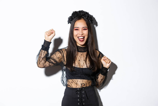 Image of young asian woman having fun on halloween party, raising hand up as rejoicing or triumphing, wearing witch gothic dress, standing over white background - Photo, Image