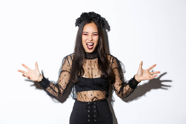Woman screaming and looking scary in her halloween witch costume, spread hands sideways and looking angry, trying scare people during trick or treat, standing over white background - Фото, изображение