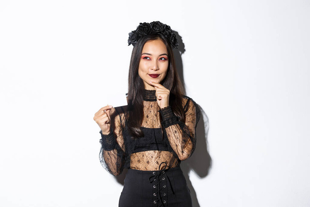 Image of smiling beautiful asian woman in gothic lace dress and wreath, thinking while holding credit card, standing over white background - Photo, Image