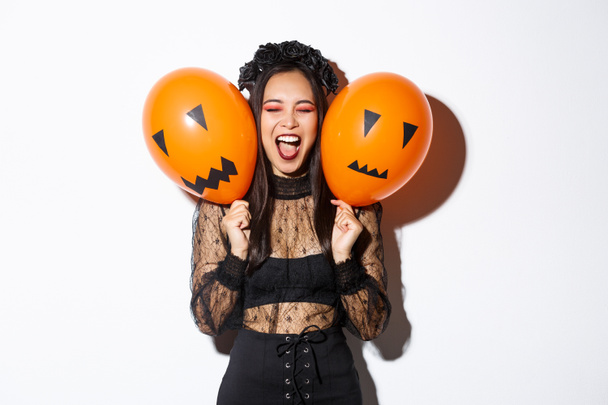 Imagen de chica asiática en traje de bruja malvada sosteniendo dos globos naranjas con caras aterradoras, celebrando halloween, de pie sobre fondo blanco - Foto, Imagen
