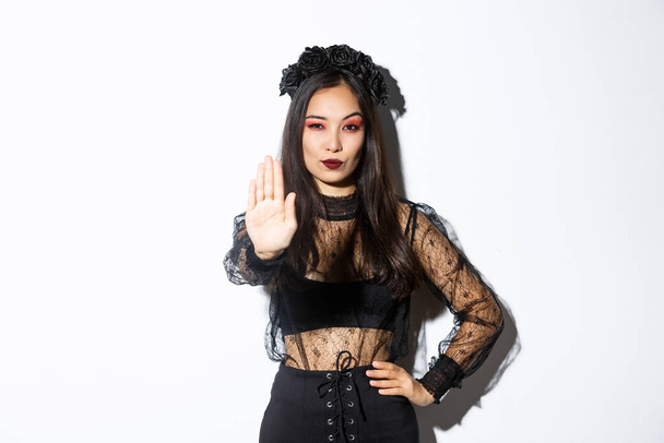 Confident elegant young witch showing stop gesture, woman dressed-up in halloween costume extend one hand to prohibit something, express disapproval, standing white background - Photo, Image