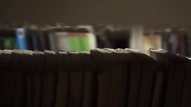 młody biały mężczyzna w różowej koszuli spaceruje w ciemnym pokoju biblioteki pomiędzy półkami z literaturą i dwoma zadbanymi czystymi rękami wyciąga jedną książkę z półki trzymając podręcznikowy palec - Materiał filmowy, wideo