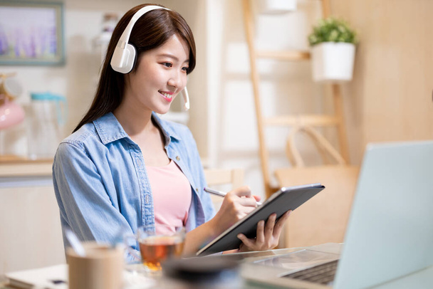 telework concept - Asian business woman uses laptop computer to join video meeting with headset and writes notes on her digital tablet at home - Photo, image