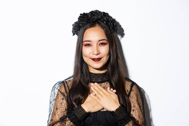 Close-up of thankful smiling asian woman looking grateful with hands over chest, standing in witch costume over white background - 写真・画像