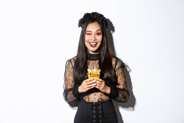 Cheeky fille souriante en costume de sorcière, célébrer Halloween, aller tour ou traiter en robe gothique, montrant la langue et tenant des bonbons - Photo, image