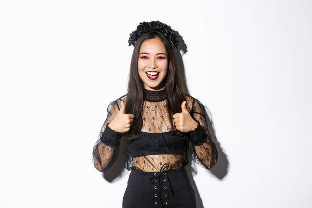 Image of happy satisfied asian woman dressed-up for halloween party, showing thumbs-up in approval, smiling pleased over white background. Girl in witch costume looking delighted - Photo, Image