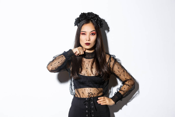 Image of beautiful gothic asian woman in black lace dress, threaten someone, make cutting throat gesture and looking serious at camera, celebrating halloween, standing over white background - Foto, Imagen