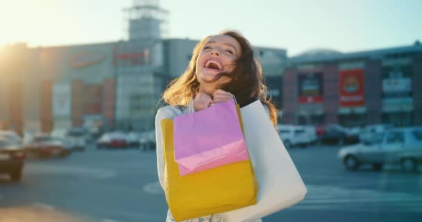 Het meisje glimlacht. Ze houdt boodschappentassen in haar handen. Ze staat op de parkeerplaats van het winkelcentrum. De zon schijnt op de achtergrond. 4K - Video