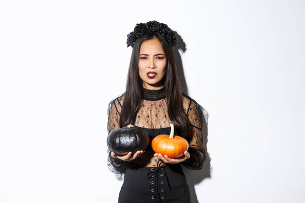 Image of grimacing angry witch throwing pumpkins, girl celebrating halloween, standing over white background - Photo, Image