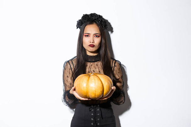 Imagen de la mujer asiática en mueca disfrazada de bruja de odio tallar calabaza para Halloween, mirando decepcionado, de pie sobre fondo blanco - Foto, imagen