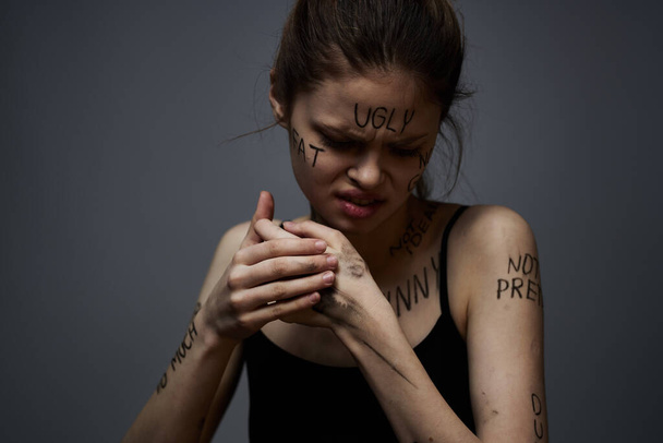 προσβεβλημένη γυναίκα με θυμωμένες επιγραφές στο σώμα της αγγίζει τον εαυτό της με τα χέρια της σε ένα γκρίζο φόντο επιθετικότητα  - Φωτογραφία, εικόνα