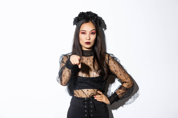 Arrogant and sassy asian evil witch, woman in halloween costume, pointing finger at you and looking mysterious, standing over white background - Photo, Image