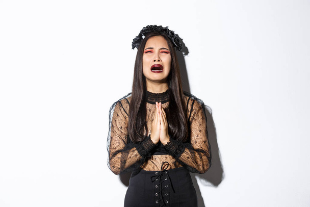 Miserable joven asiática mujer en problemas suplicando a Dios, llorando y pidiendo ayuda, usando vestido gótico de halloween y corona, suplicando sobre fondo blanco - Foto, imagen
