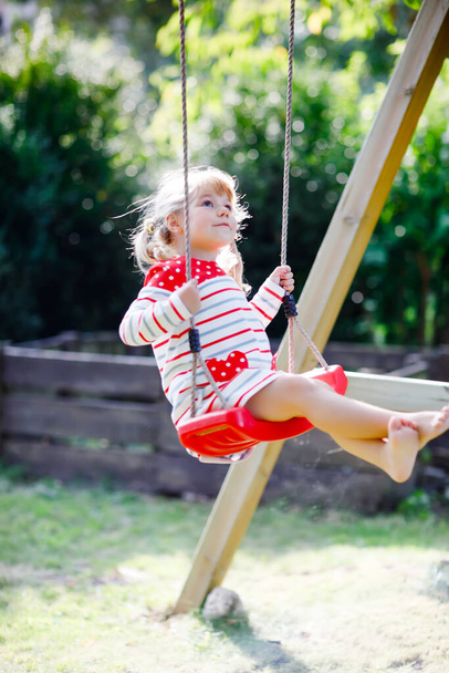 Szczęśliwa piękna dziewczynka maluch zabawy na huśtawce w ogrodzie. Słodkie zdrowe dziecko huśtawki pod kwitnącymi drzewami w słoneczny wiosenny dzień. Dziecko śmieje się i płacze - Zdjęcie, obraz
