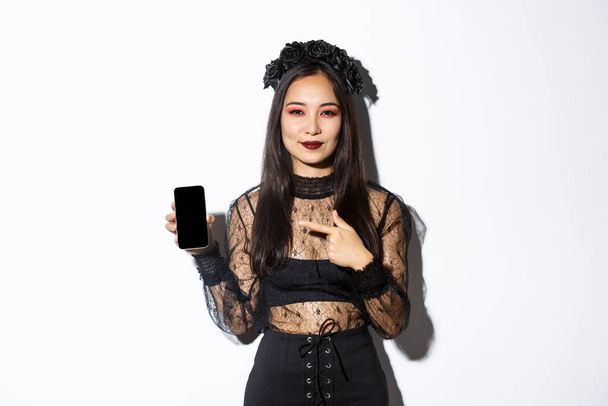Jonge elegante vrouw in gotische jurk en zwarte krans wijzen vinger naar smartphone scherm met een tevreden glimlach op haar gezicht, staande over witte achtergrond - Foto, afbeelding