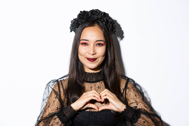 Detailní záběr usmívající se krásné asijské ženy v kostýmu čarodějnice a černý věnec ukazující srdce gesto, milující Halloween dovolená, stojící nad bílým pozadím - Fotografie, Obrázek