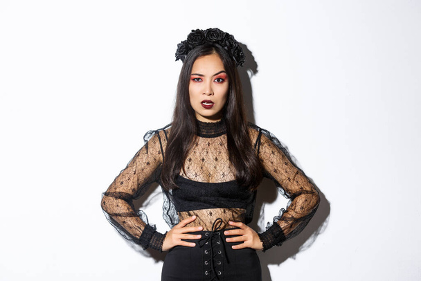 Angry and pissed-off asian female magician, evil witch in black dress and wreath looking mad at someone, frowning and looking judgemental, standing over white background - Photo, image