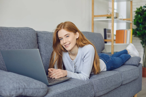 Smiling girl lying on sofa with laptop and looking at camera at home - Fotoğraf, Görsel