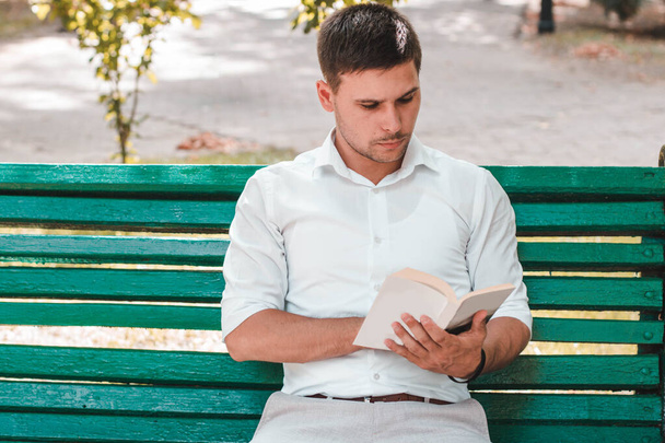 Молодой человек в белой рубашке читает книгу в парке - Фото, изображение