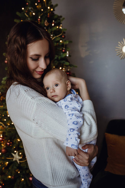 Maman en pull blanc embrasse le petit garçon le gamin dans le pull au sapin de Noël et les guirlandes dans la maison - Photo, image