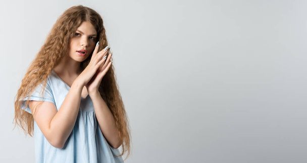 Photo of thoughtful European young woman with curly long hair looks with dreamy expression aside, stands against white wall with free space for text - Photo, Image