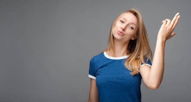 Funny young blonde female shoots in temple, tilts head, dressed in casual blue t shirt, demonstrates suicide gesture, isolated on gray background with blank space - Photo, Image
