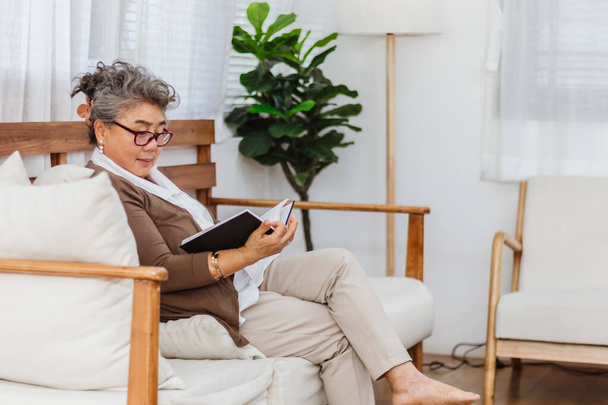 Boldog és egészségesen nyugalmazott mosolygós idős nő vagy nyugdíjas olvasó könyvet pihen, vagy ül a kanapén vagy kanapén otthon. Idős felnőtt aktív életmód és aktivitás a nyaralás. Egészség- és biztosítási koncepció - Fotó, kép