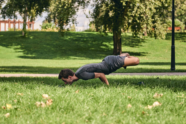 Молодой спортсмен в спортивной одежде занимается йогой в парке. Практика asana на открытом воздухе. Люди тренируются на зеленой траве с ковриком для йоги. Сильный взрослый кавказский мужчина в сложной позе павлина лотоса. - Фото, изображение