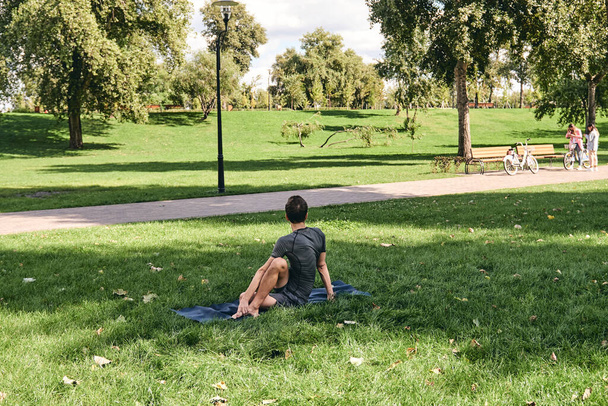 Mladý atletický muž ve sportovním oblečení dělá jógu v parku. Trénujte půl páteře zvrátit asana venku. Lidé cvičí na zelené trávě s podložkou na jógu. Fitness a zdravý životní styl. - Fotografie, Obrázek