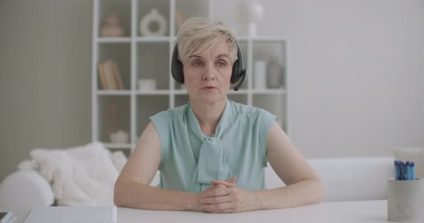 ηλικιωμένη γυναίκα μιλάει σε ακουστικά με μικρόφωνο και κοιτάζοντας την κάμερα, κάθεται στο τραπέζι στο σπίτι, call-center και τεχνολογία τηλεδιάσκεψης - Πλάνα, βίντεο