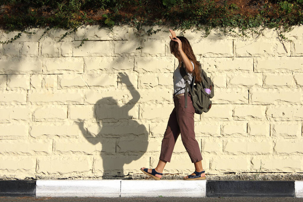 Joven turista con mochila detrás de su espalda se divierte, jugando con la sombra, caminando lentamente a lo largo de la acera a lo largo del camino - Foto, Imagen