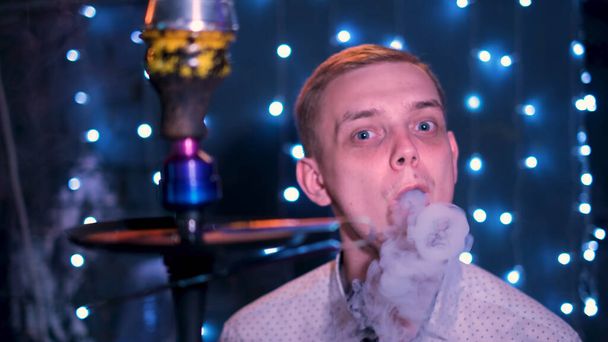 Jeune homme blond aux yeux bleus soufflant des anneaux de fumée. Les médias. Homme fumant narguilé au bar de nuit, expirant fumée sur fond de lumières bleues brillantes. - Photo, image