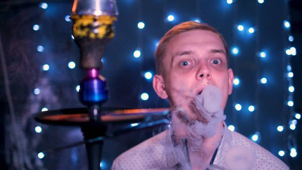 Young blond man with blue eyes blowing smoke rings. Media. Male smoking hookah at the night bar, exhaling smoke on blue shining lights background. - Valokuva, kuva